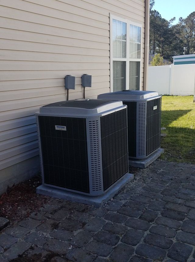 Two gray outdoor HVAC units beside a beige house on a stone patio near a grassy yard