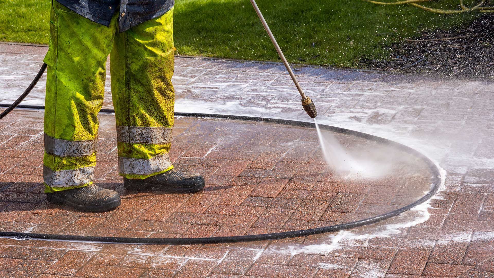 Worker pressure-washing a wet pavement, with spray nozzle and green-yellow safety pants visible
