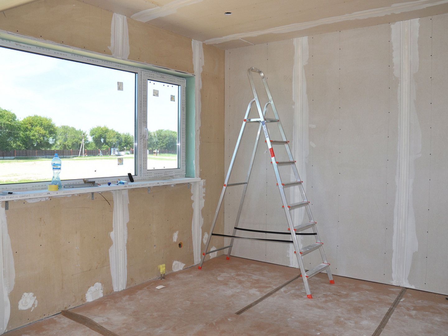 Interior room under renovation with a ladder beside a large window and unfinished walls.
