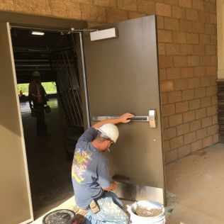 Person kneels by a metal door in a hallway, using a paint roller on the handle area.