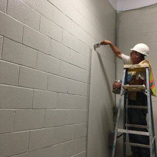 Worker on a ladder painting a cinder block wall in a gray hallway