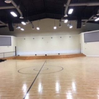 Empty indoor gymnasium with wooden floor, center court circle, stage at one end, and overhead lights