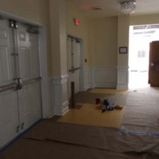 Dim hallway with double doors, white wainscoting, and a tarp-covered floor with tools near a bright doorway.