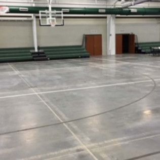 Empty indoor gym with polished floor, basketball hoop, and folded green bleachers along the wall