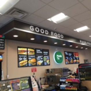 Convenience store counter with “GOOD FOOD” sign, illuminated menu boards, and a drinks display
