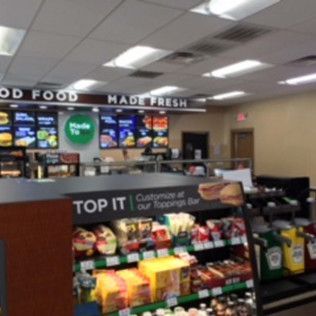 Convenience store checkout with snack displays and menu boards under bright fluorescent lights