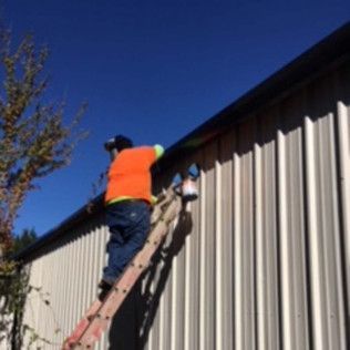 Worker in orange vest climbs a ladder to repair a metal-sided house roof under a blue sky