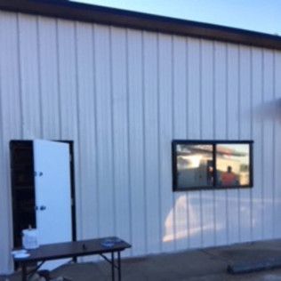 Exterior of a white metal building with an open door, window, and folding table outside