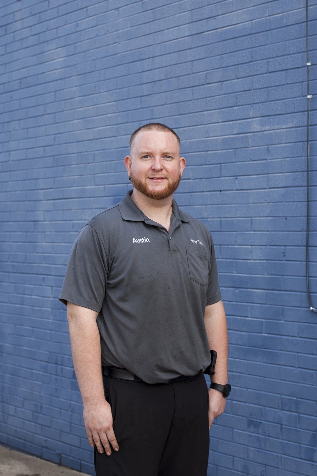 Man standing against a blue brick wall, wearing a gray polo shirt and black pants.