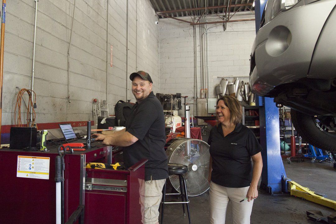Two people standing beside a vehicle in a garage, with a car lift and shop equipment nearby.