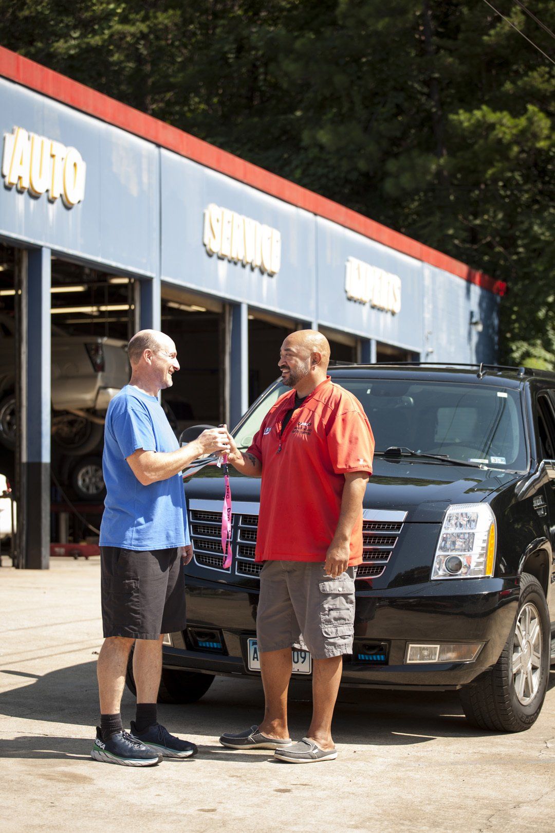 Two men talking beside a black SUV outside an auto shop.