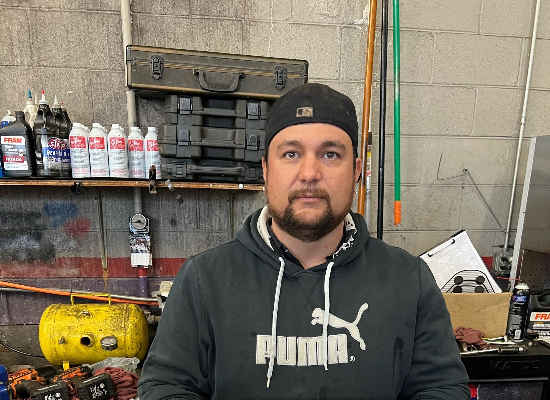 Man in black Puma hoodie and cap standing in a workshop with tools and supplies behind him