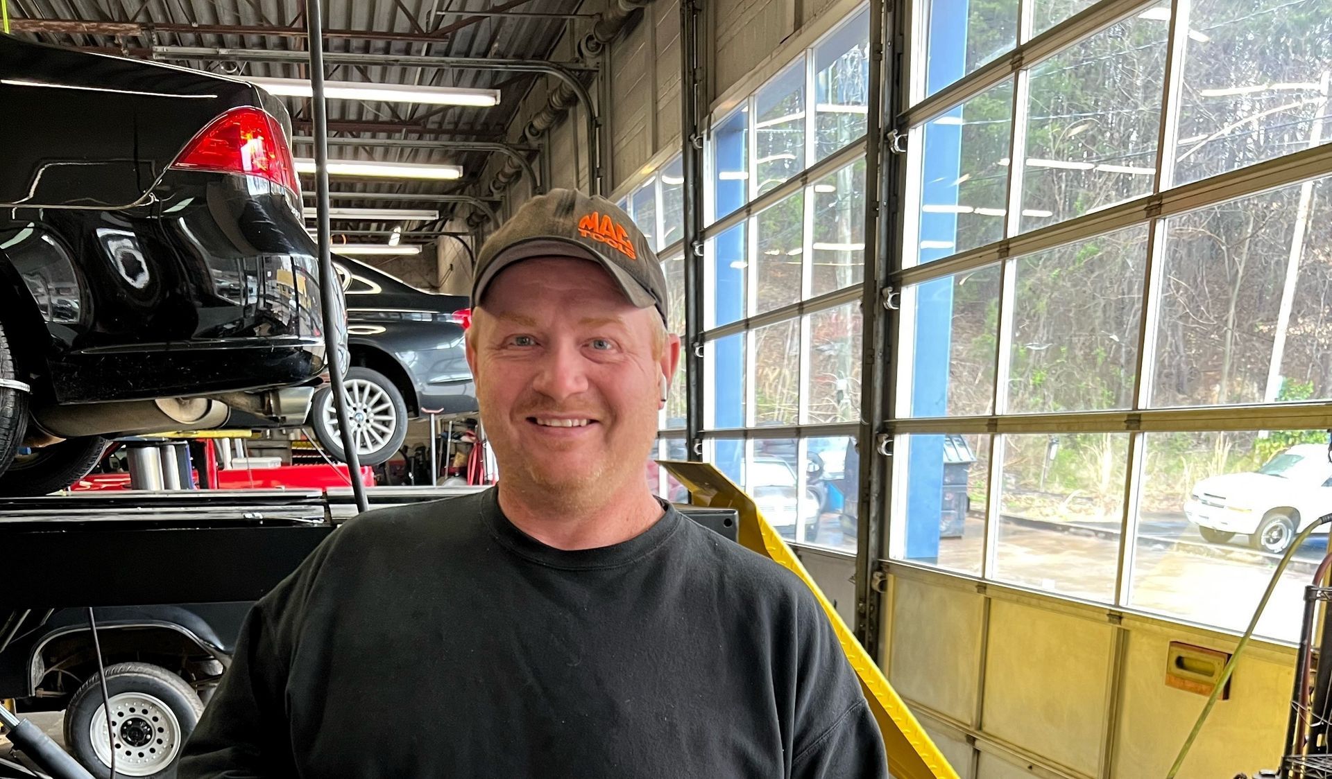 Man smiling in a workshop beside stacked cars, with a yellow-railed windowed wall in the background