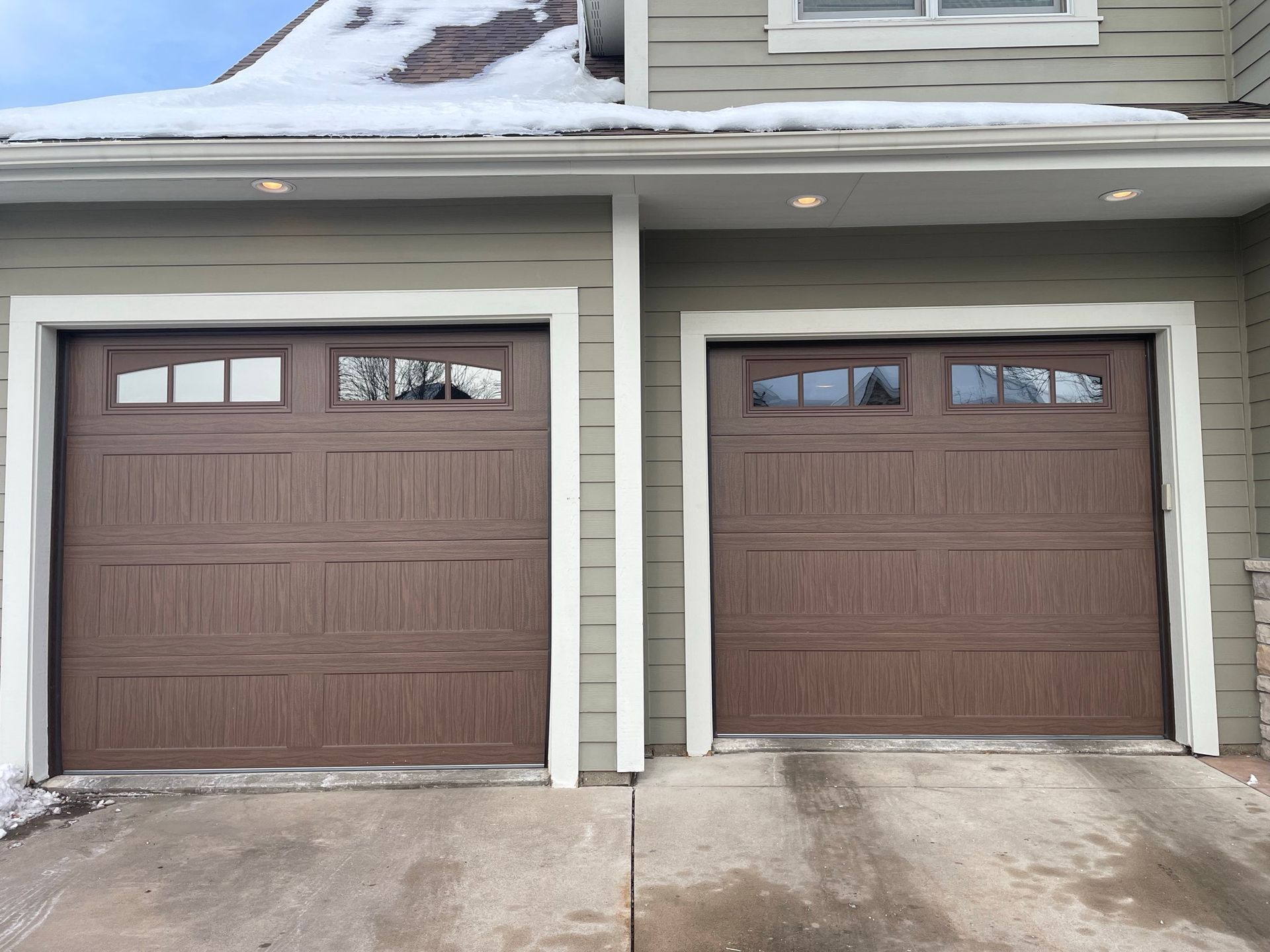 Two brown garage doors with white trim on a house, with snow on the roof.