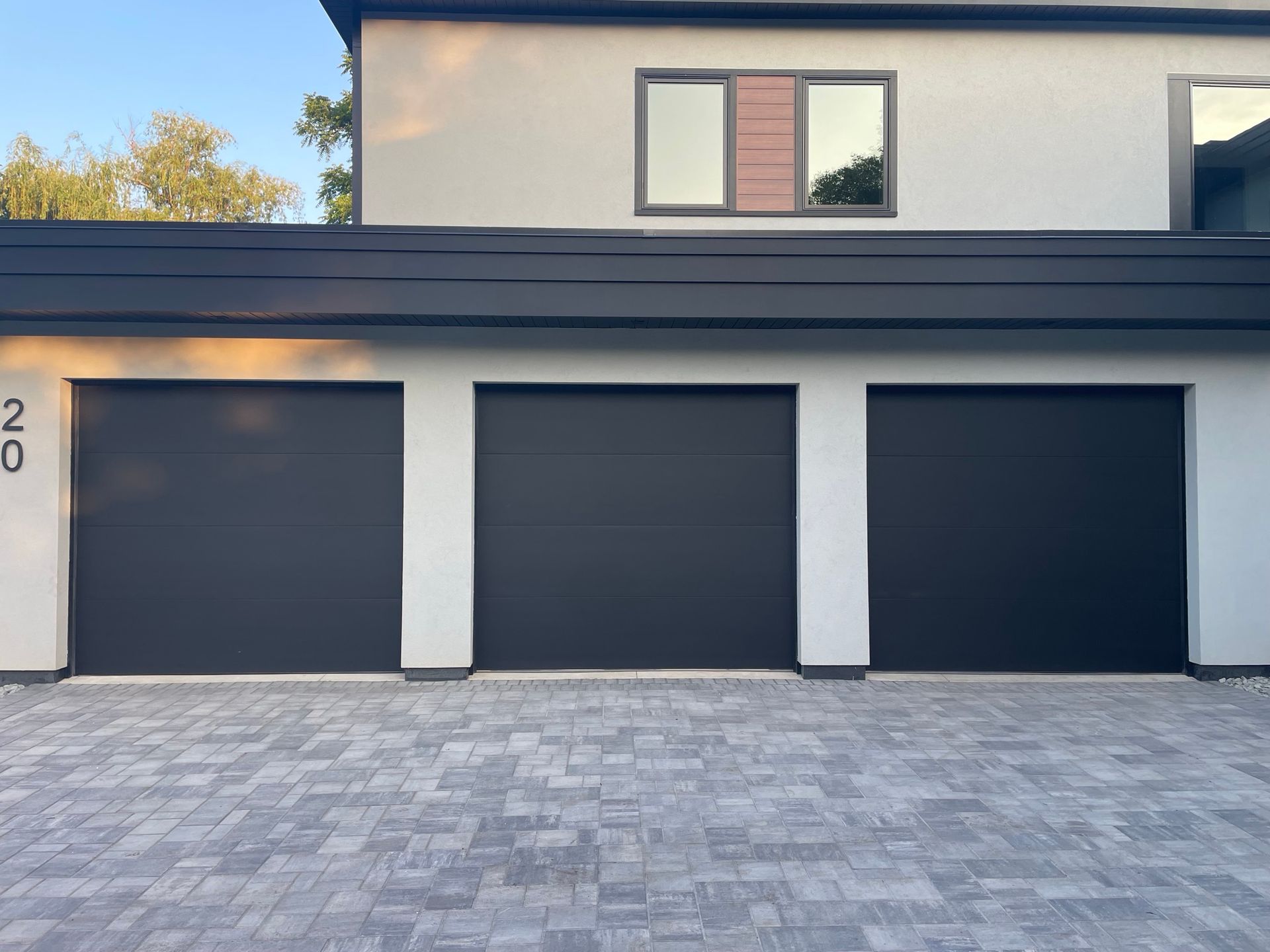 Three black garage doors on a light-colored building, grey brick driveway, dark roof.