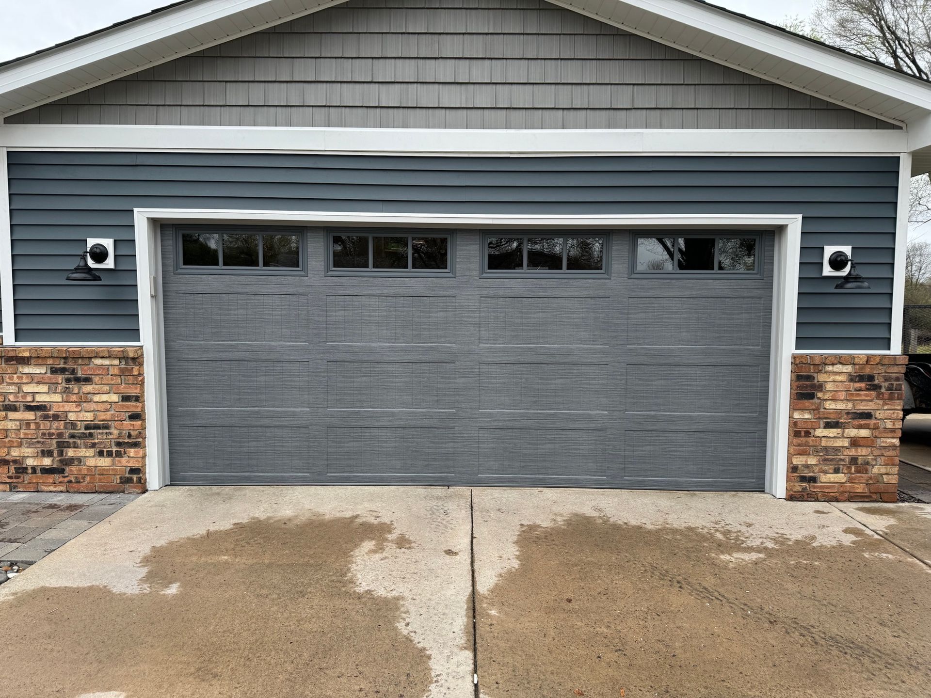Gray garage door with window panels, blue siding, brick accents. Concrete driveway.