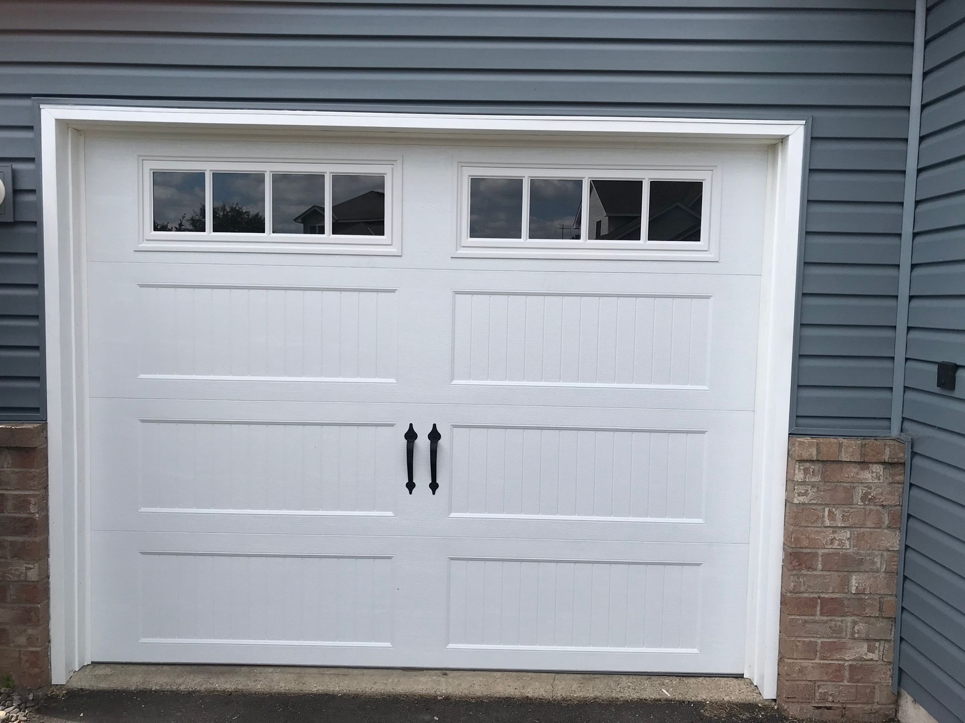 White garage door with window panels and black handles, set against blue siding and brick.