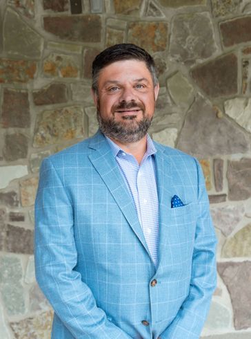 Man in a light blue blazer standing in front of a stone wall, smiling