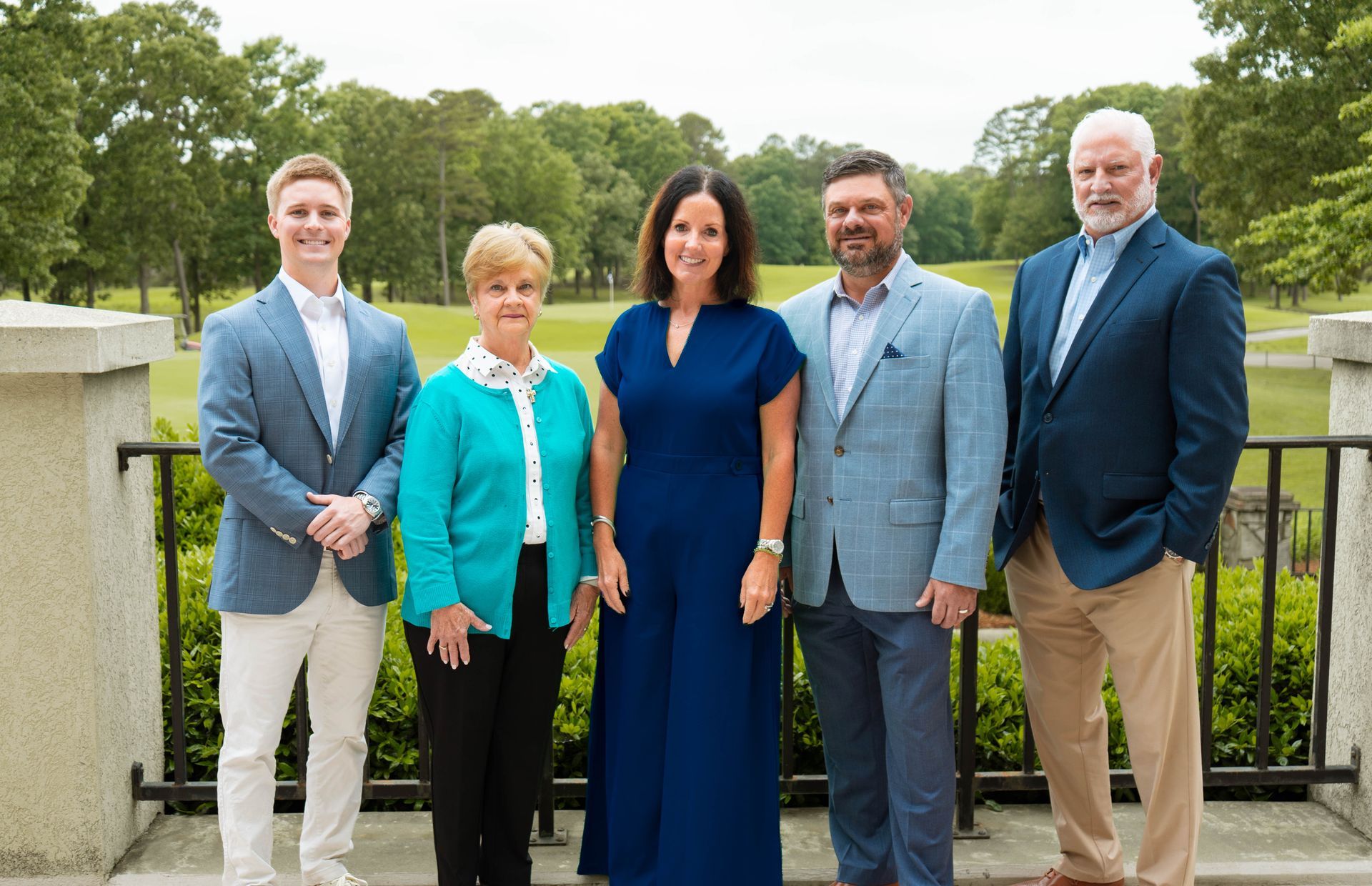 Five people standing outdoors in a park, dressed in formal and business attire.