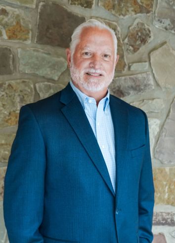Man in a blue blazer standing against a stone wall, smiling at the camera.