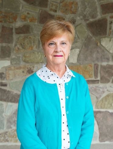 Woman standing against a stone wall, wearing a turquoise cardigan and white polka-dot blouse.