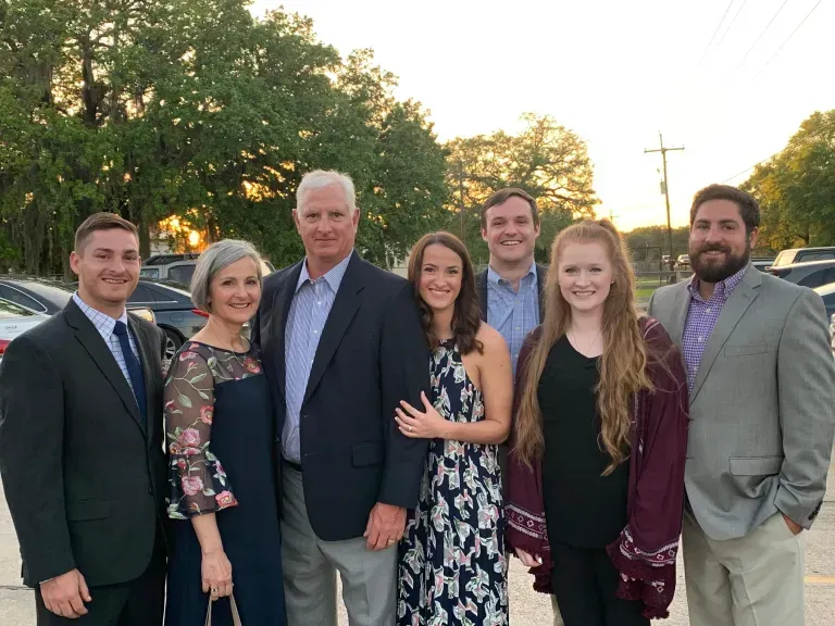 Seven people posing outdoors at sunset, dressed formally, smiling near trees and a road.