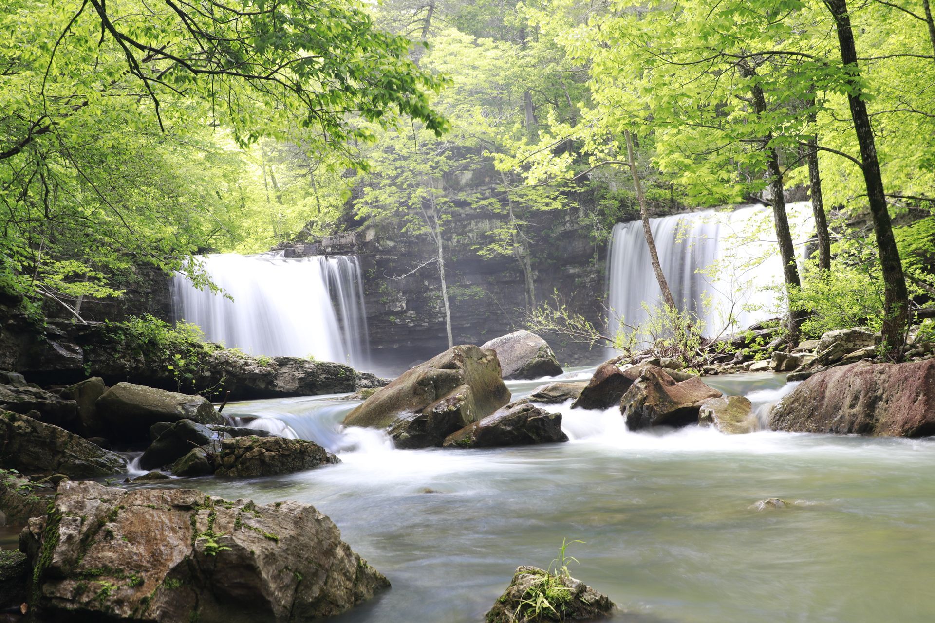 Forest waterfall cascading into a rocky river, framed by green trees and mist.