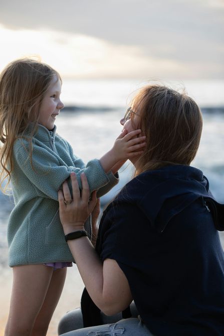 Person kneels on a beach, gently holding a child’s face at sunset.