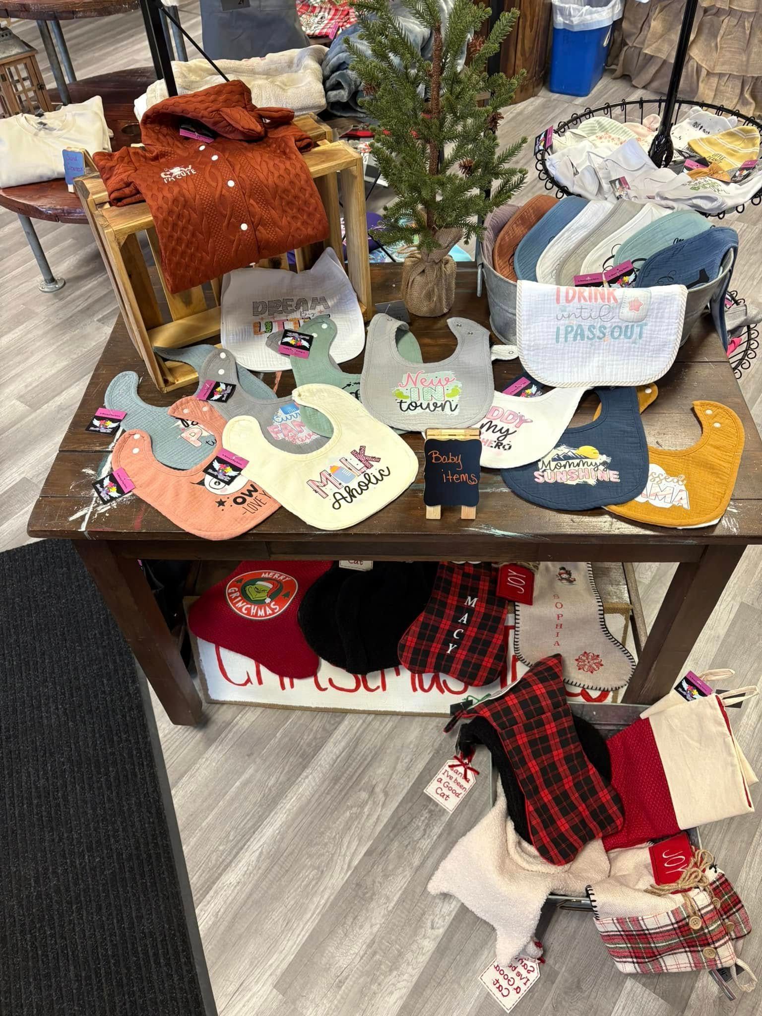 A wooden store display table filled with assorted baby bibs, a small decorative tree, and Christmas stockings below.