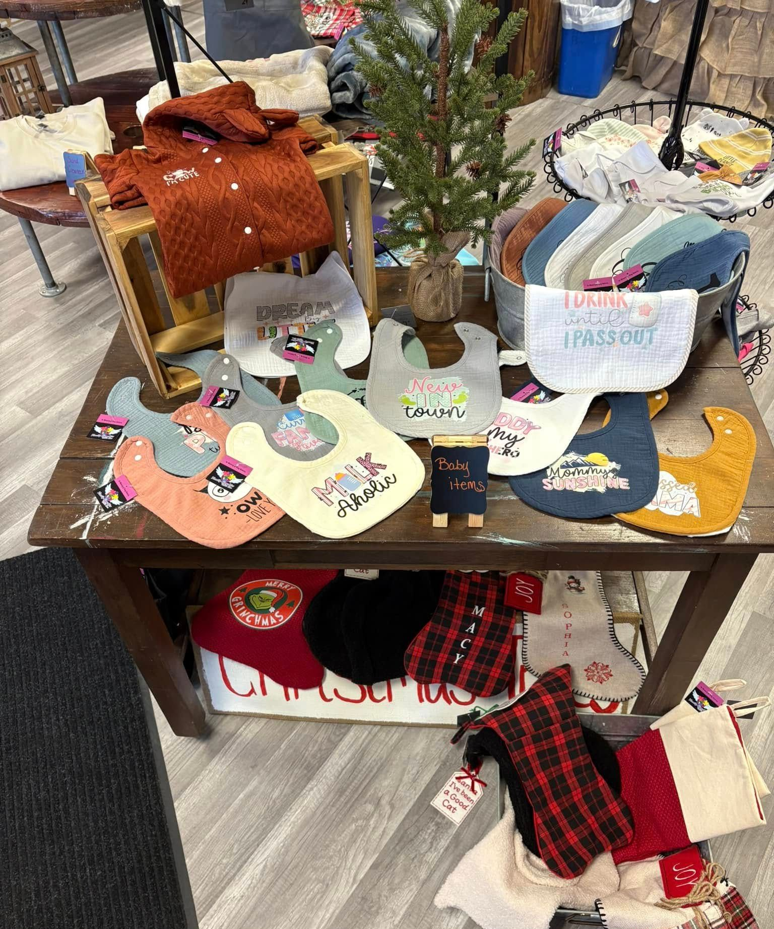 Display table featuring baby bibs, a small evergreen tree, and a knit sweater, with Christmas stockings placed below.