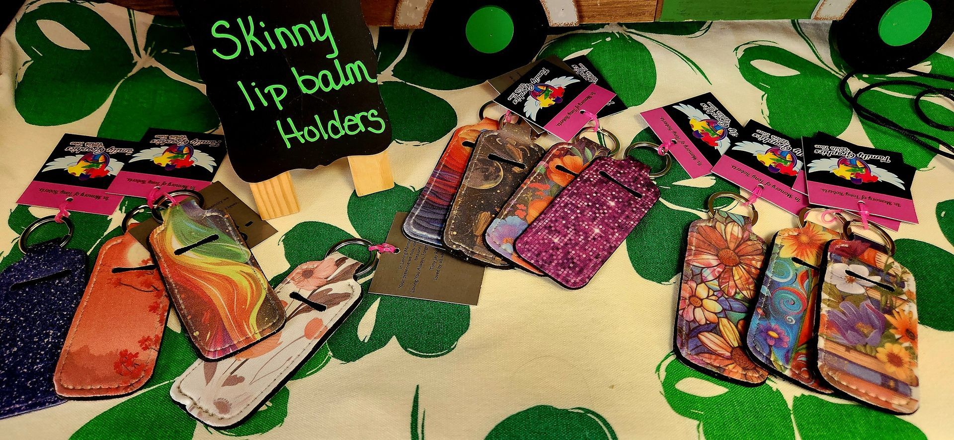 Assorted skinny lip balm holders with various patterns displayed on a green clover tablecloth with a small wooden sign.