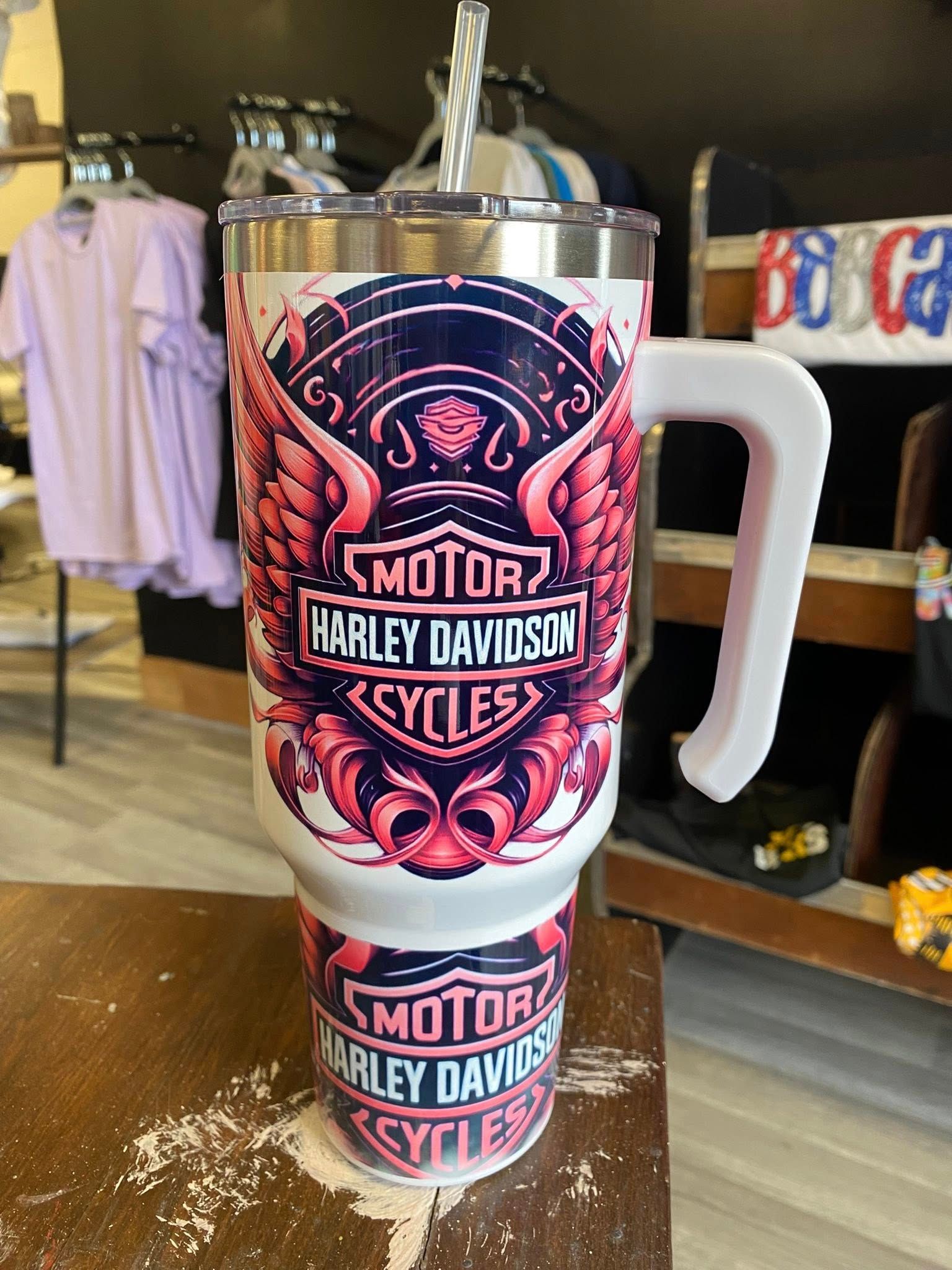 A white Harley-Davidson tumbler with a large pink-winged logo, featuring a handle and straw, sitting on a wooden table.
