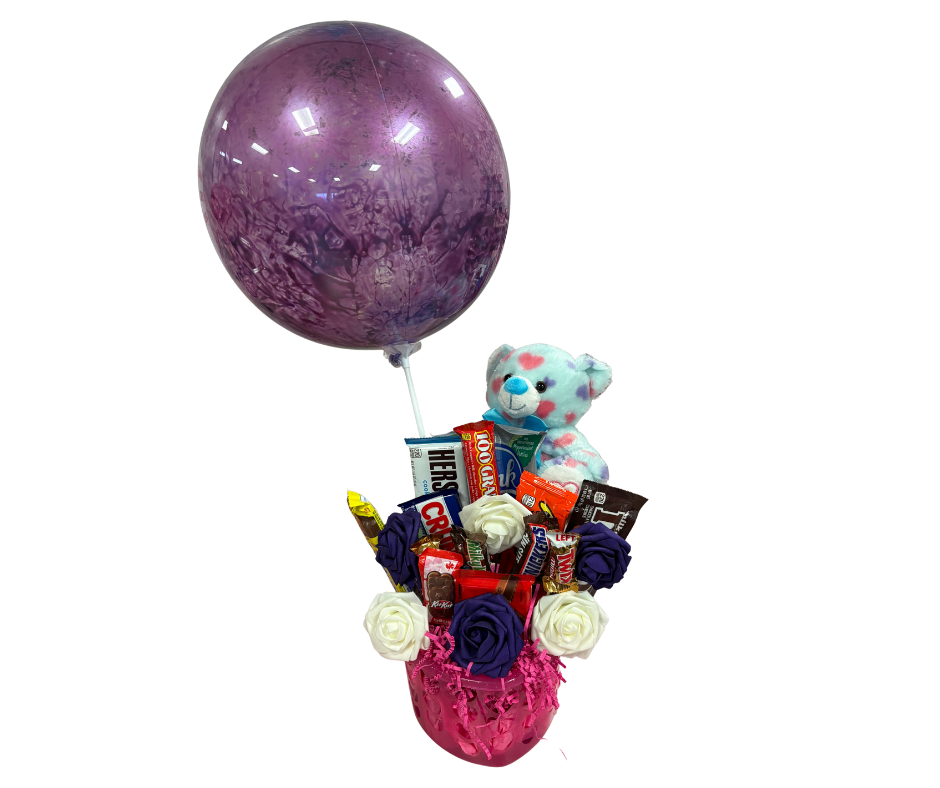 A gift arrangement featuring a purple balloon, a small blue bear, assorted candy bars, and fabric flowers in a pink base.