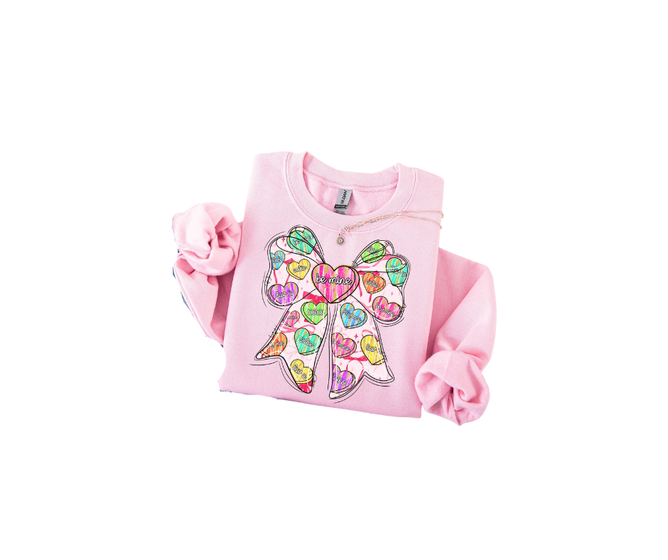 A light pink sweatshirt featuring a large graphic of a bow decorated with colorful conversation heart candies.
