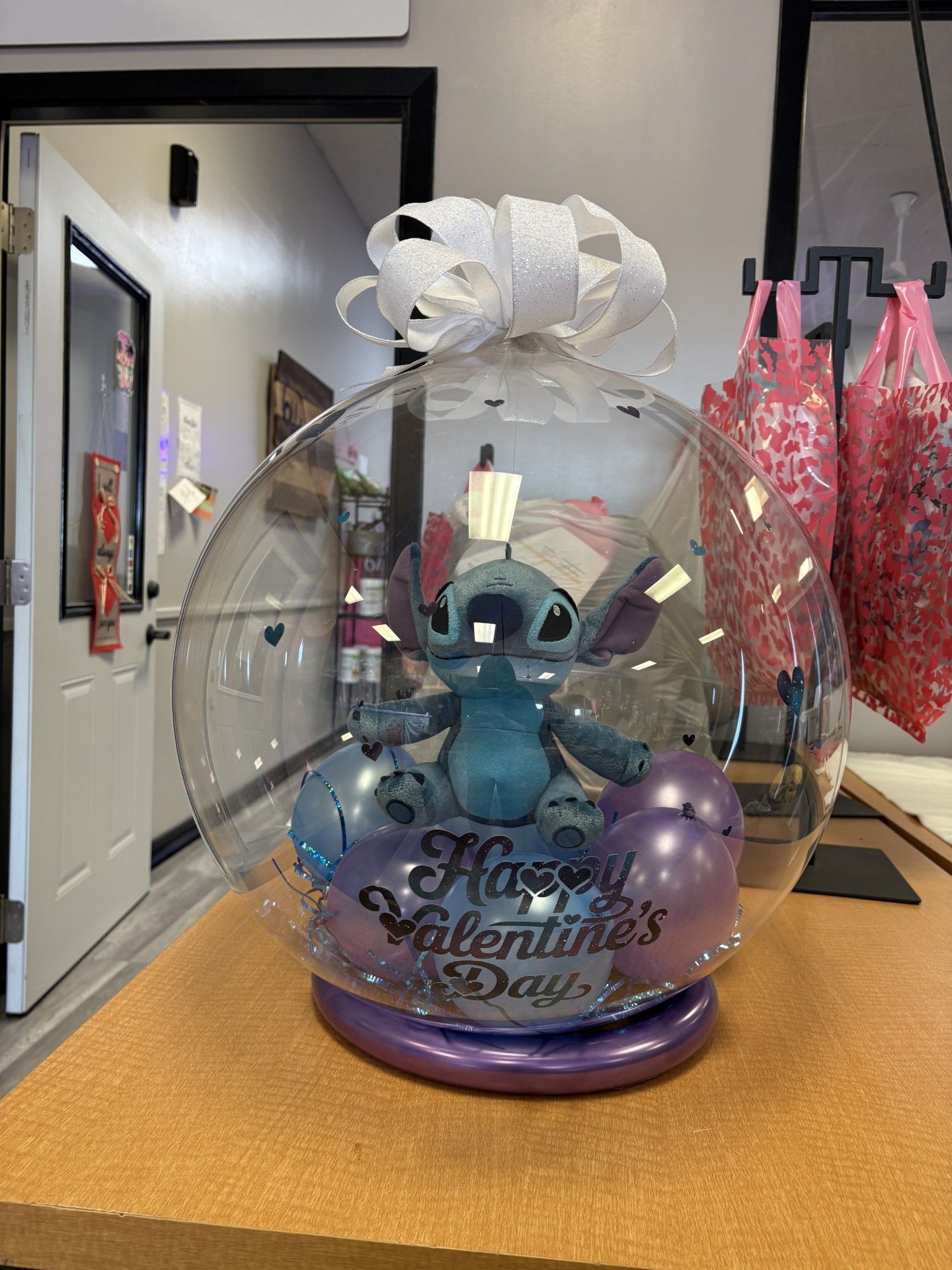 A Stitch plush toy sits inside a clear, spherical balloon filled with small purple balloons, marked 