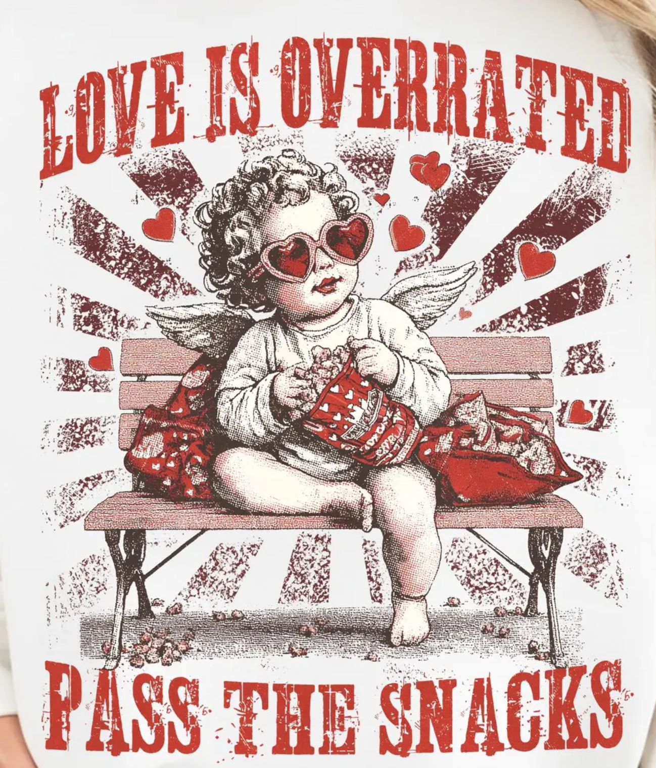 A cupid wearing heart-shaped sunglasses sits on a park bench eating snacks, with text: 