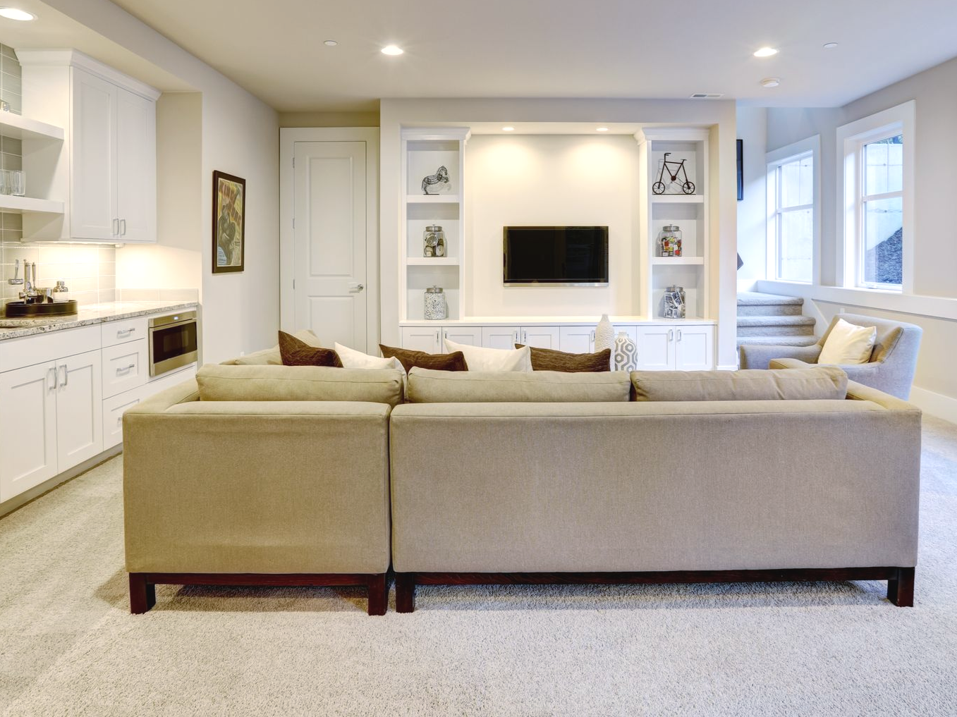 A living room with a large beige sectional sofa facing a built-in media center with a mounted television and white shelves.