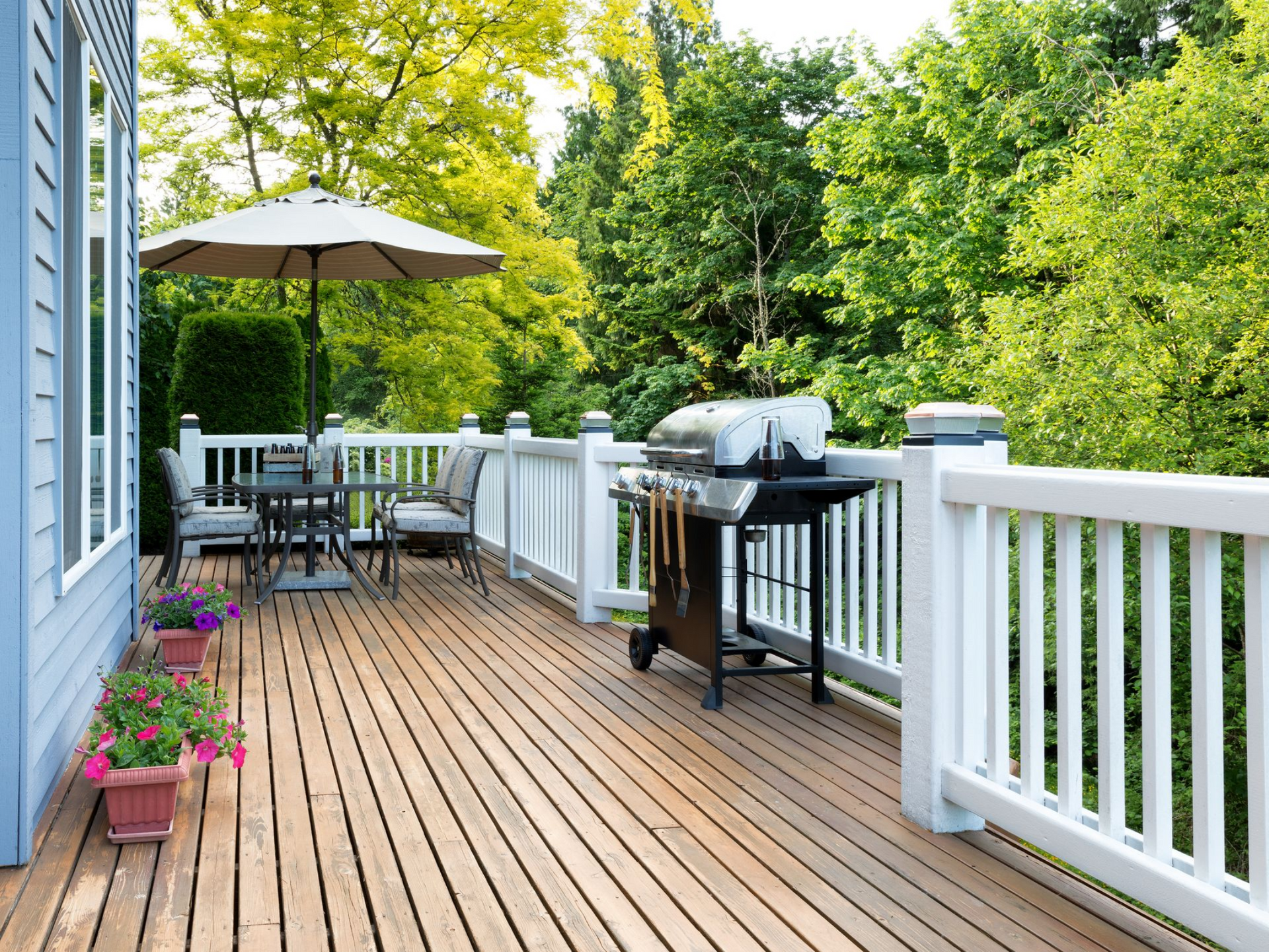 A wooden deck featuring a dining table with an umbrella, a grill, and potted flowers against a backdrop of green trees.