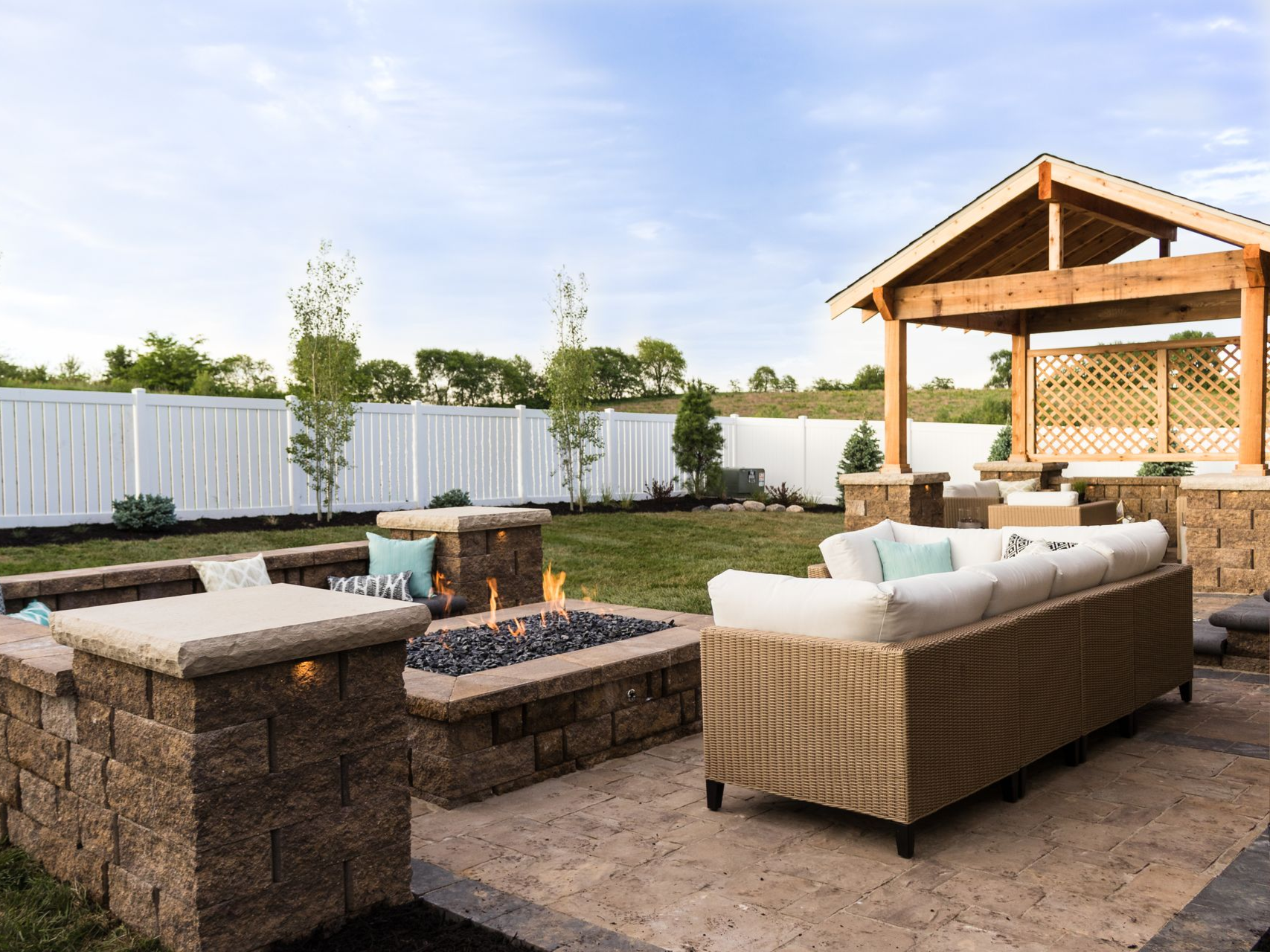 A patio with a stone fire pit, beige wicker sofa, and a wooden gazebo in a backyard with a white fence.