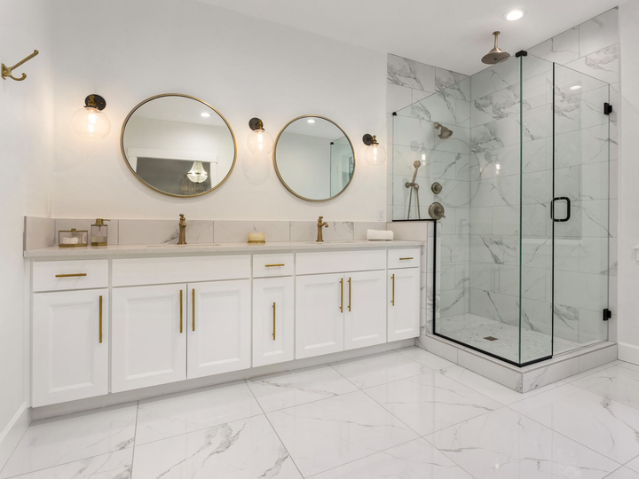 Modern bathroom with double vanity, white cabinets, gold fixtures, circular mirrors, and a glass-enclosed marble shower.