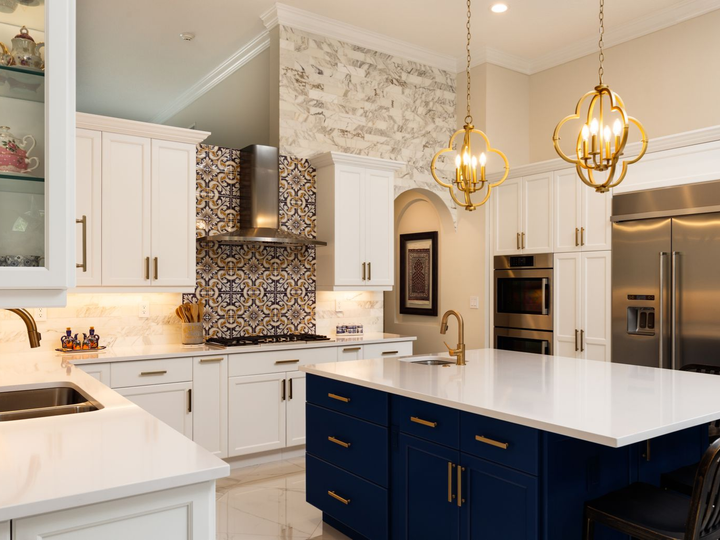 Modern kitchen featuring white cabinets, a navy blue island, white countertops, ornate wall tile, and gold pendant lights.