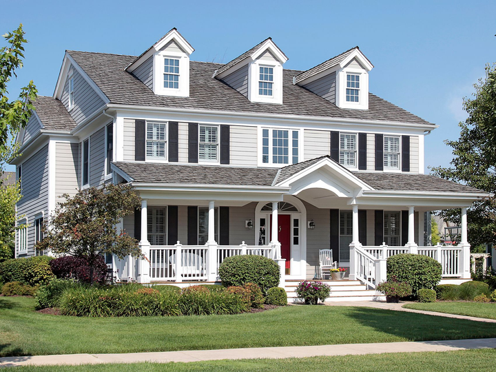 A two-story grey suburban house with three dormers, a front porch, and a white fence, surrounded by a lawn and landscaping.