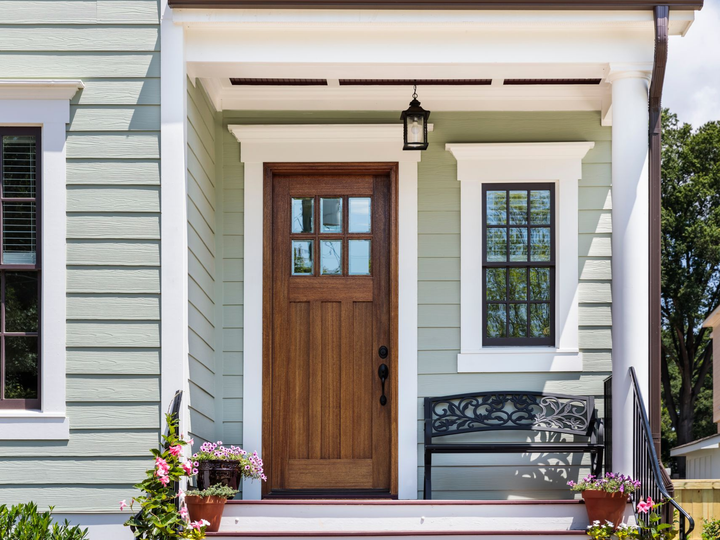 A wooden front door with six window panes on a porch, featuring sage green siding, white trim, and a black metal bench.