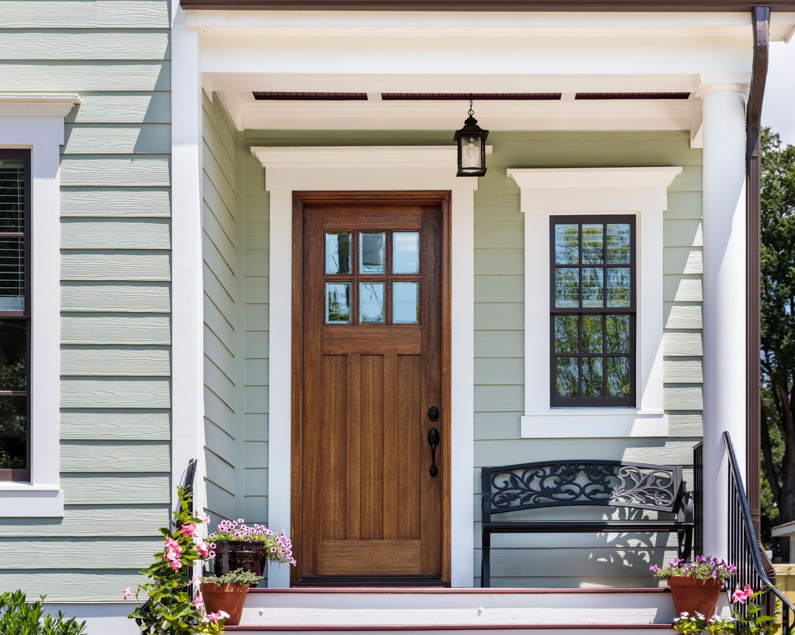 Front porch of a house with sage green siding, a wooden front door with a window, a window, and a metal bench.