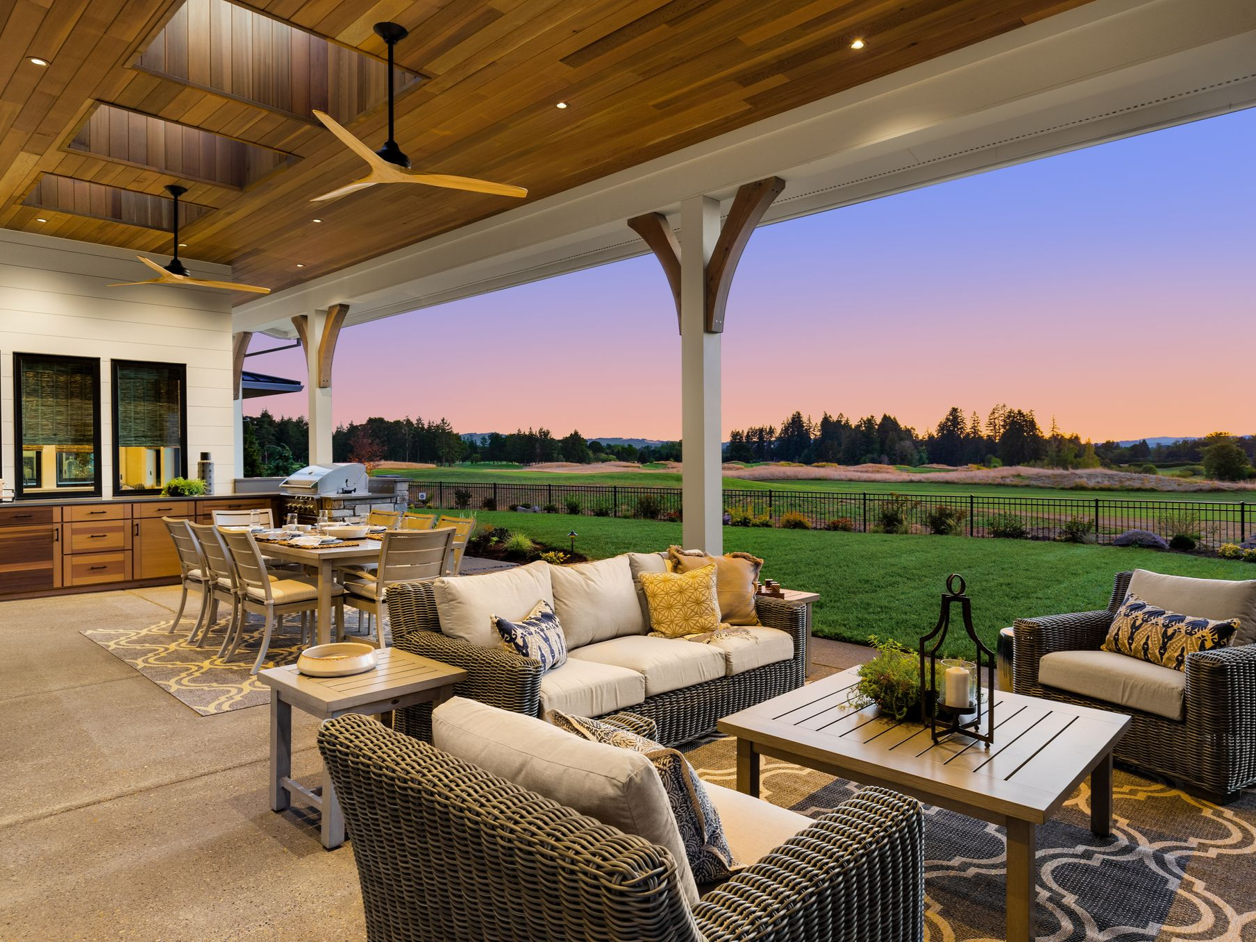 A luxury outdoor patio with wicker furniture, a dining table, and wood-paneled ceiling, overlooking a grassy field at dusk.