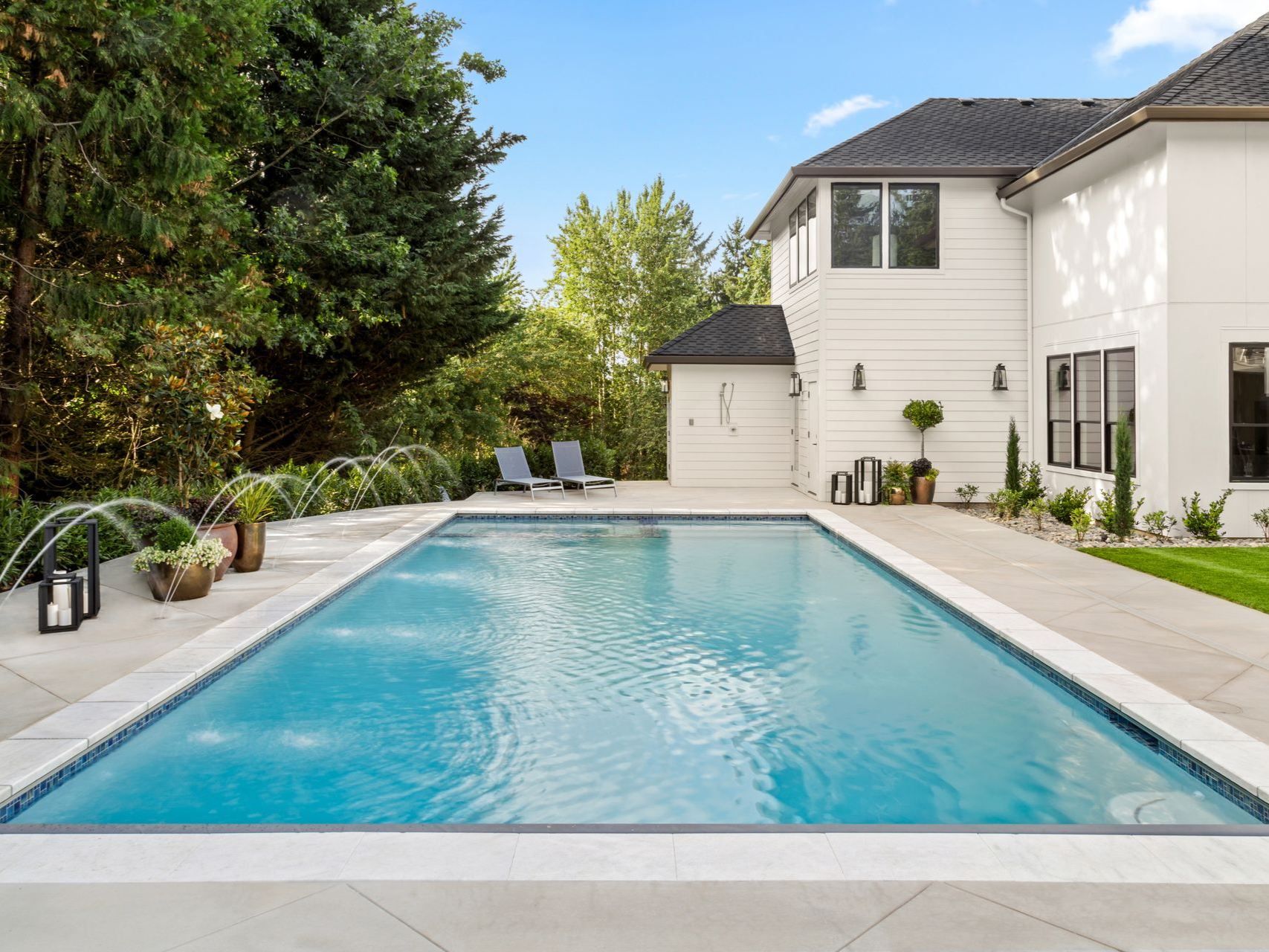 A rectangular backyard swimming pool with water fountains, surrounded by light stone decking and a white two-story house.