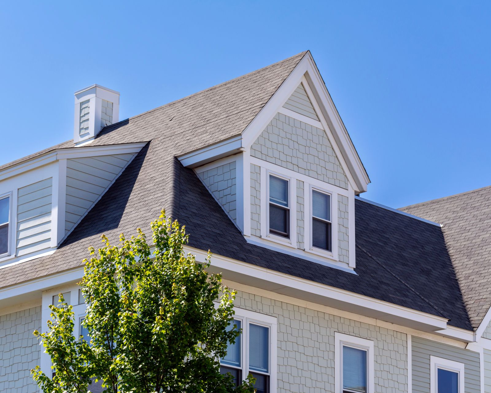 A pale green house with a shingled roof, dormer windows, and a chimney against a clear blue sky.