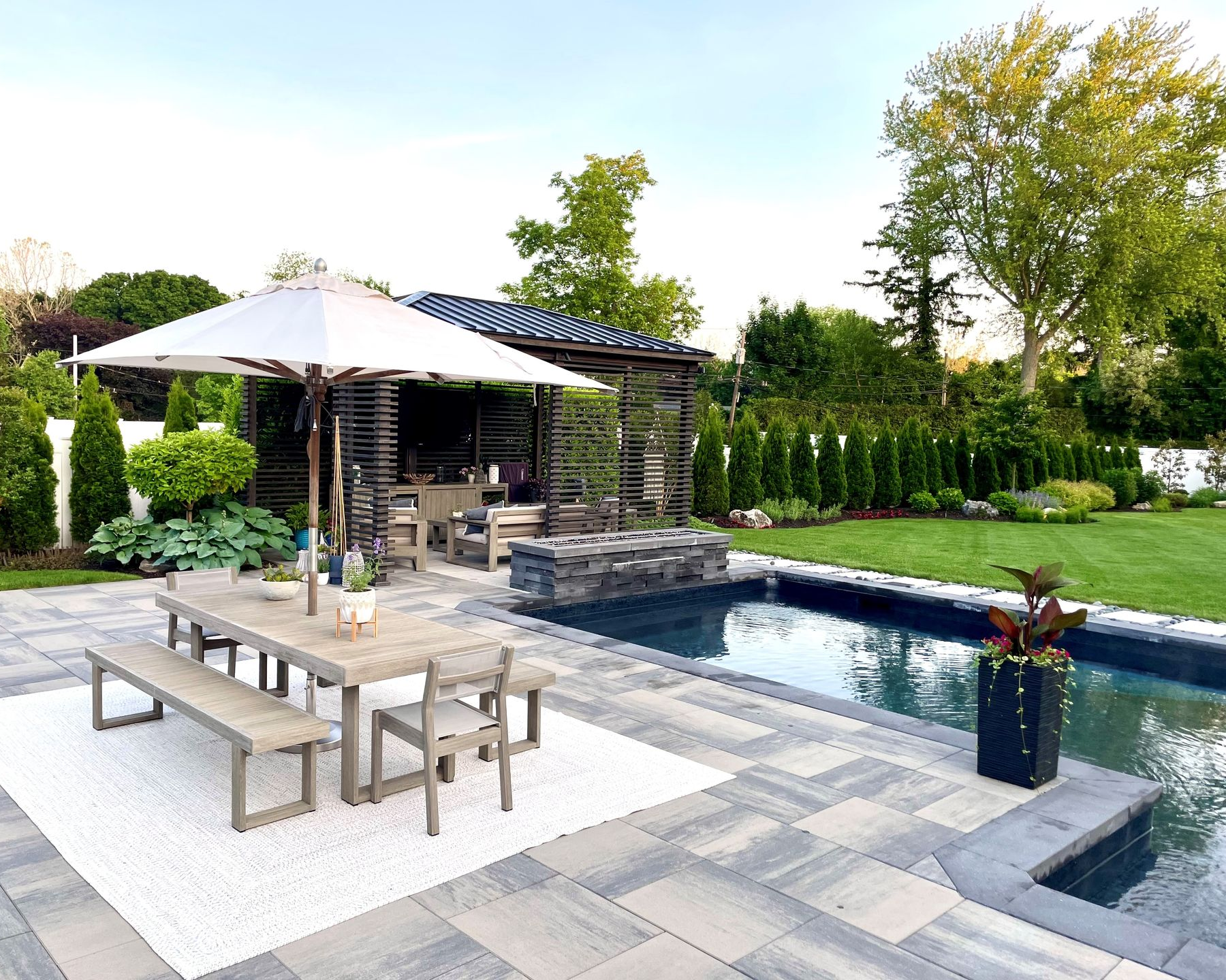 A patio with a wooden dining set under a large umbrella, beside a swimming pool and an open-air gazebo in a garden.