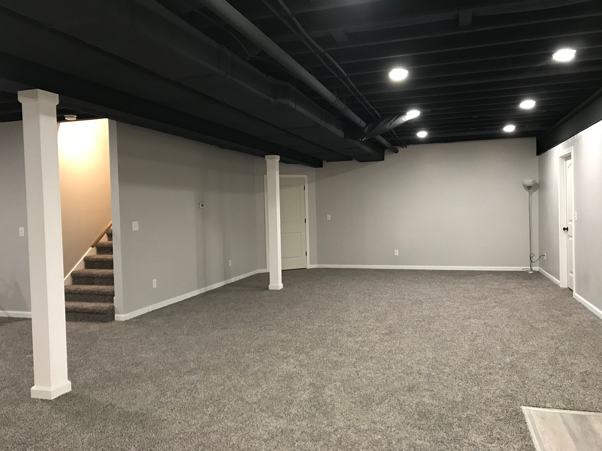 Empty finished basement room with gray carpet, white walls, exposed black ceiling beams, and a staircase.