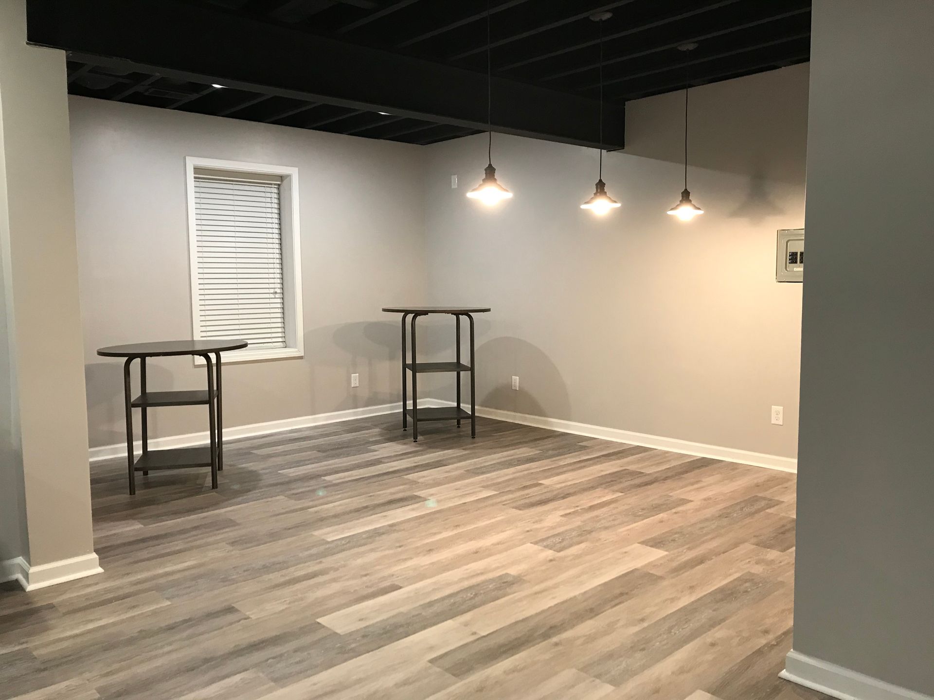 Empty basement room with gray walls, wood-look floor, and two small tables under hanging lights